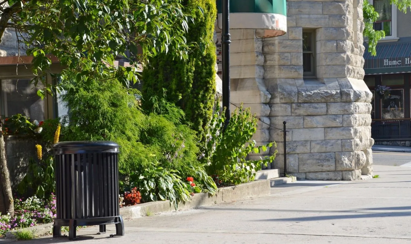 A Beselt Square Top Waste Receptacle stands on a sidewalk next to a lush garden bed. The black powder-coated bin complements the surrounding greenery and stone architecture in Nelson, British Columbia.