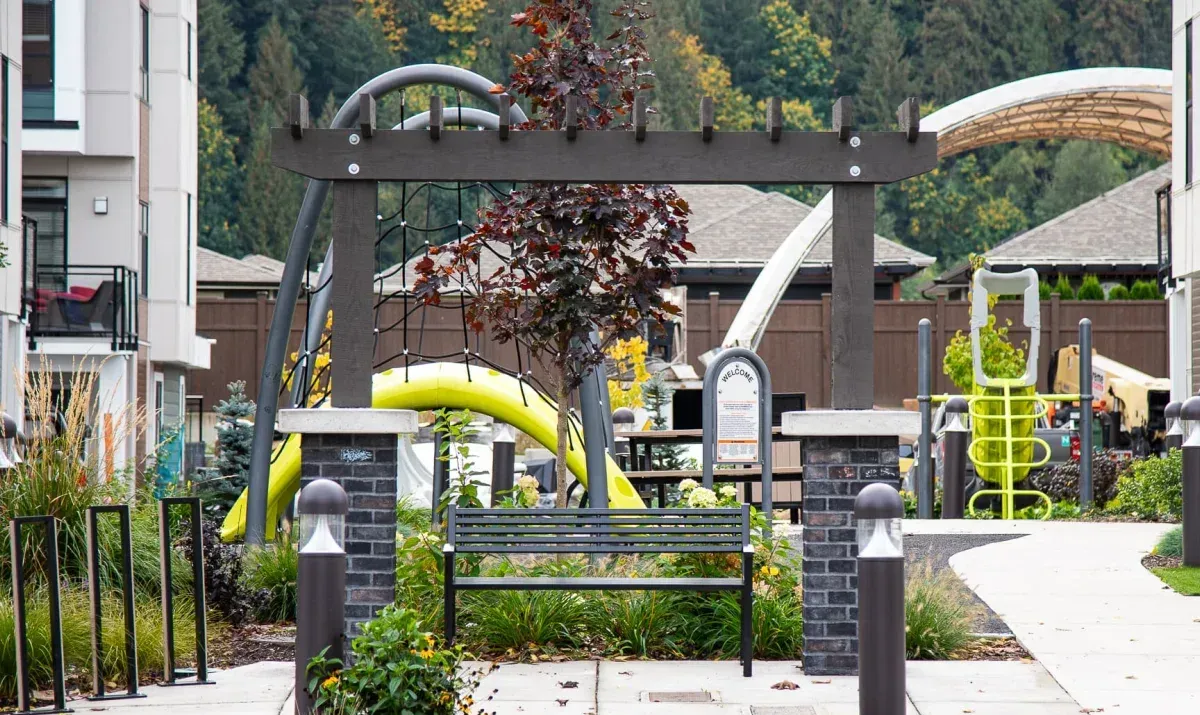 A Beselt bench provides seating in a landscaped area with a playground in the background. The bench features a black frame and horizontal slats.