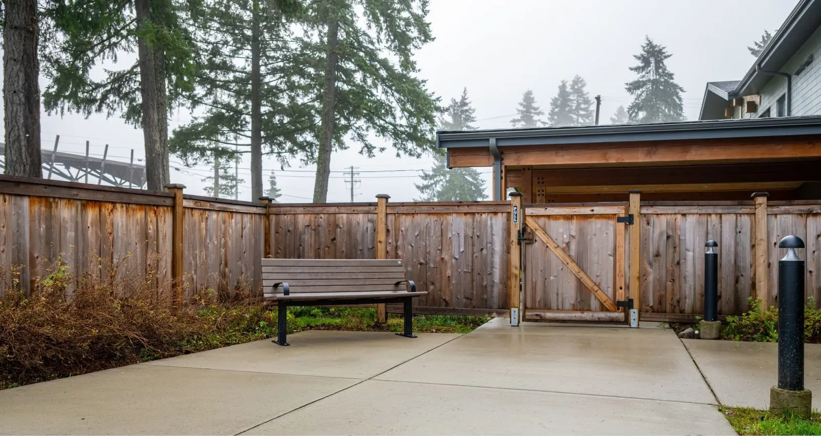 A Chat Bench provides seating in an outdoor area with a wooden fence and gate. The bench features recycled plastic lumber and a black powder-coated frame.
