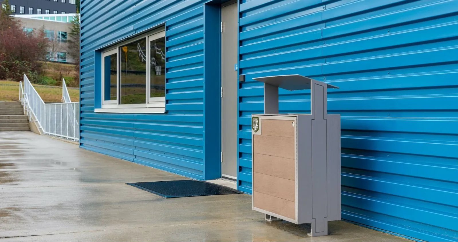 A City Scape waste receptacle stands outside a blue building at Vancouver Island University. The bin features recycled plastic lumber and a powder-coated metal frame.