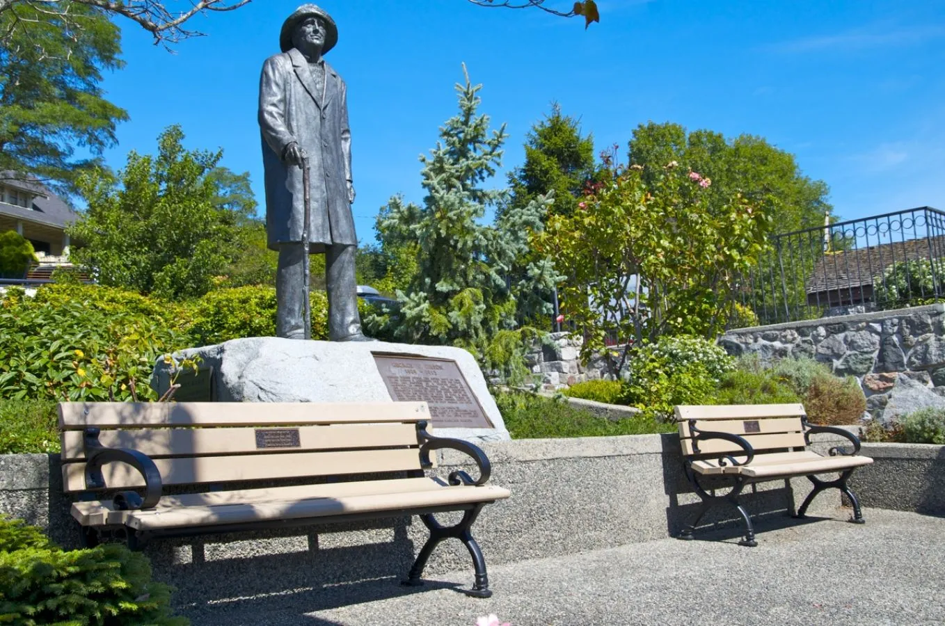 Two benches are situated in a park-like setting near a statue. The benches feature a light-colored recycled plastic lumber and black powder-coated frames.