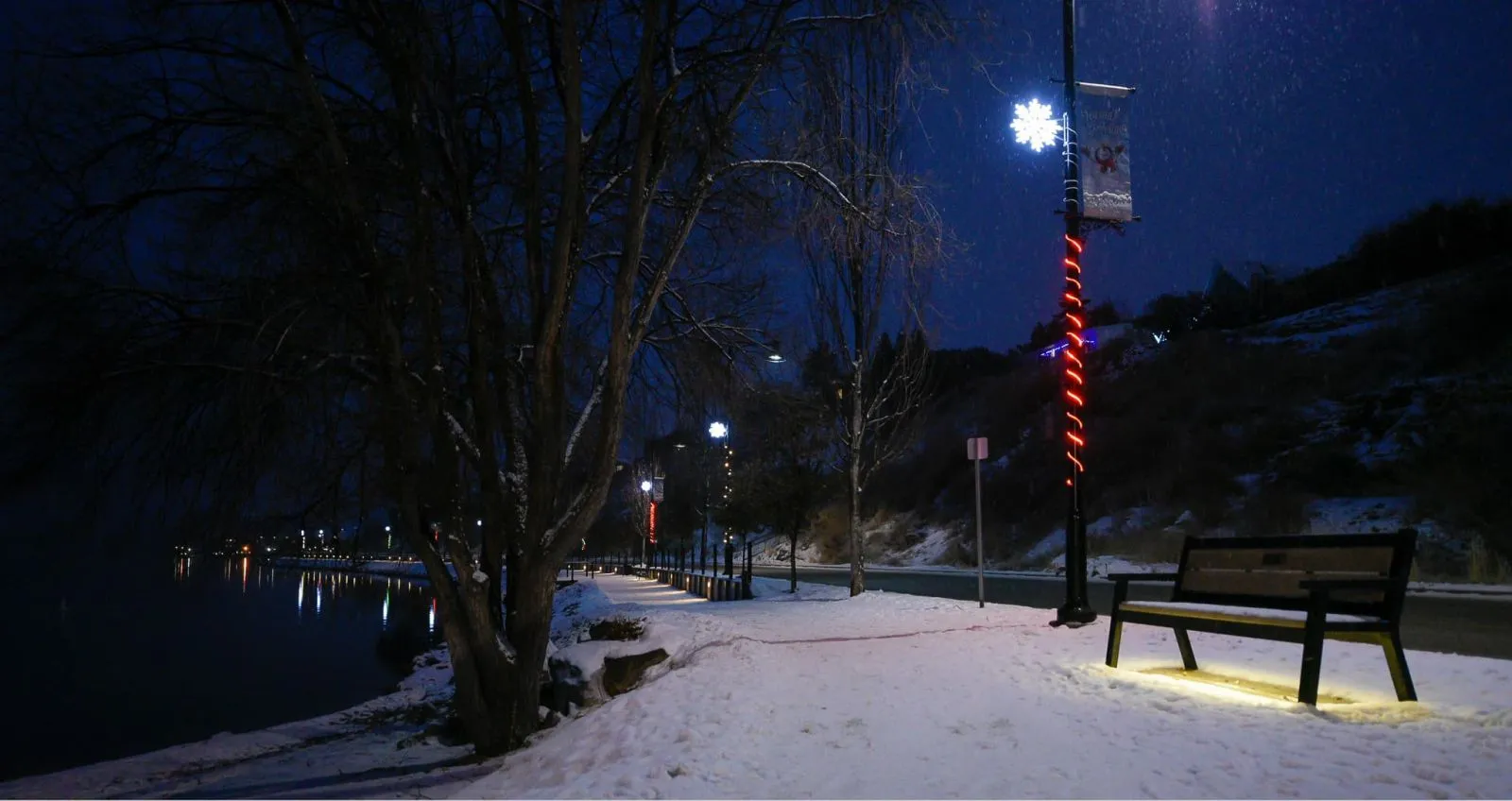A Rutherford Bench sits along a snow-covered waterfront path at night, illuminated by nearby streetlights. The bench provides a place to rest and enjoy the view of the water.