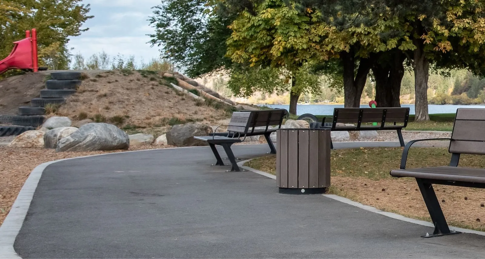 Benches and a Pipeline Waste Receptacle are installed along a paved path in a park setting. The benches feature recycled plastic lumber and black powder-coated frames.