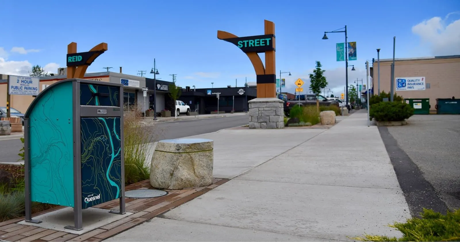 Animal-resistant waste receptacle featuring a custom graphic wrap is installed on a downtown street. The receptacle has a grey frame and a teal and navy blue graphic.