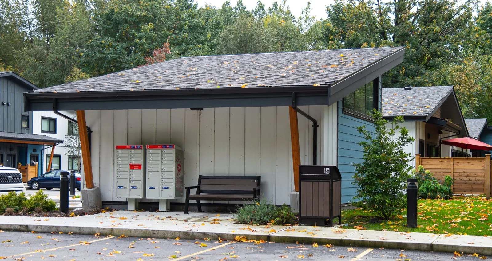 A Freedom 55 Bear Resistant Waste Receptacle and a bench sit outside a building. This durable waste bin is designed to withstand bear interactions, making it ideal for parks and public spaces.