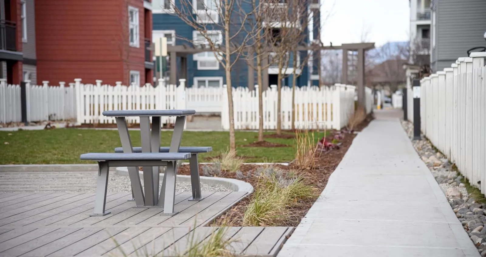A picnic table with benches sits on a wooden deck in a residential area. The table features a light gray powder-coated frame and gray recycled plastic lumber.