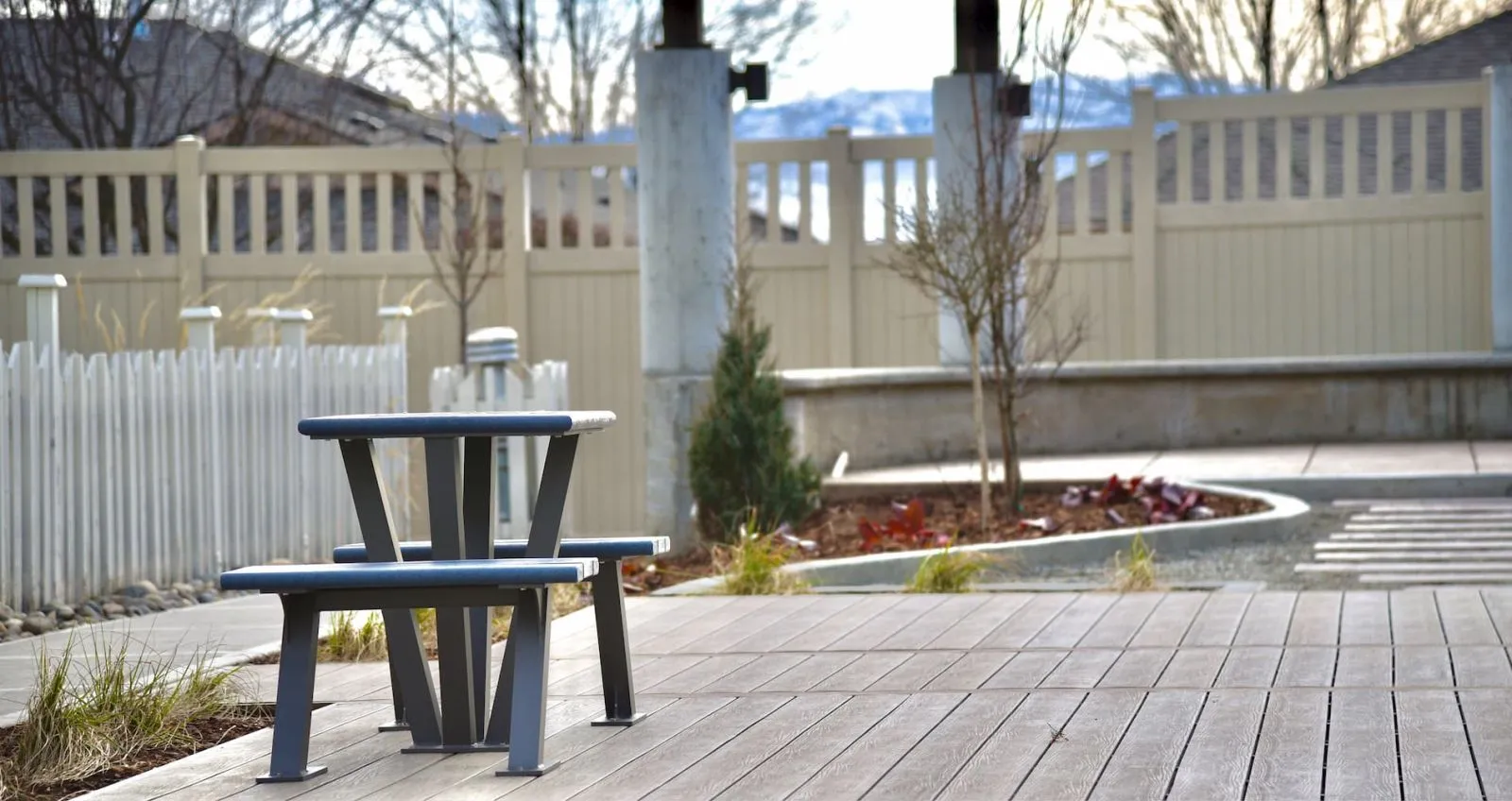 A games table sits on a wooden deck in an outdoor space. The surrounding area features a decorative fence and landscaping.