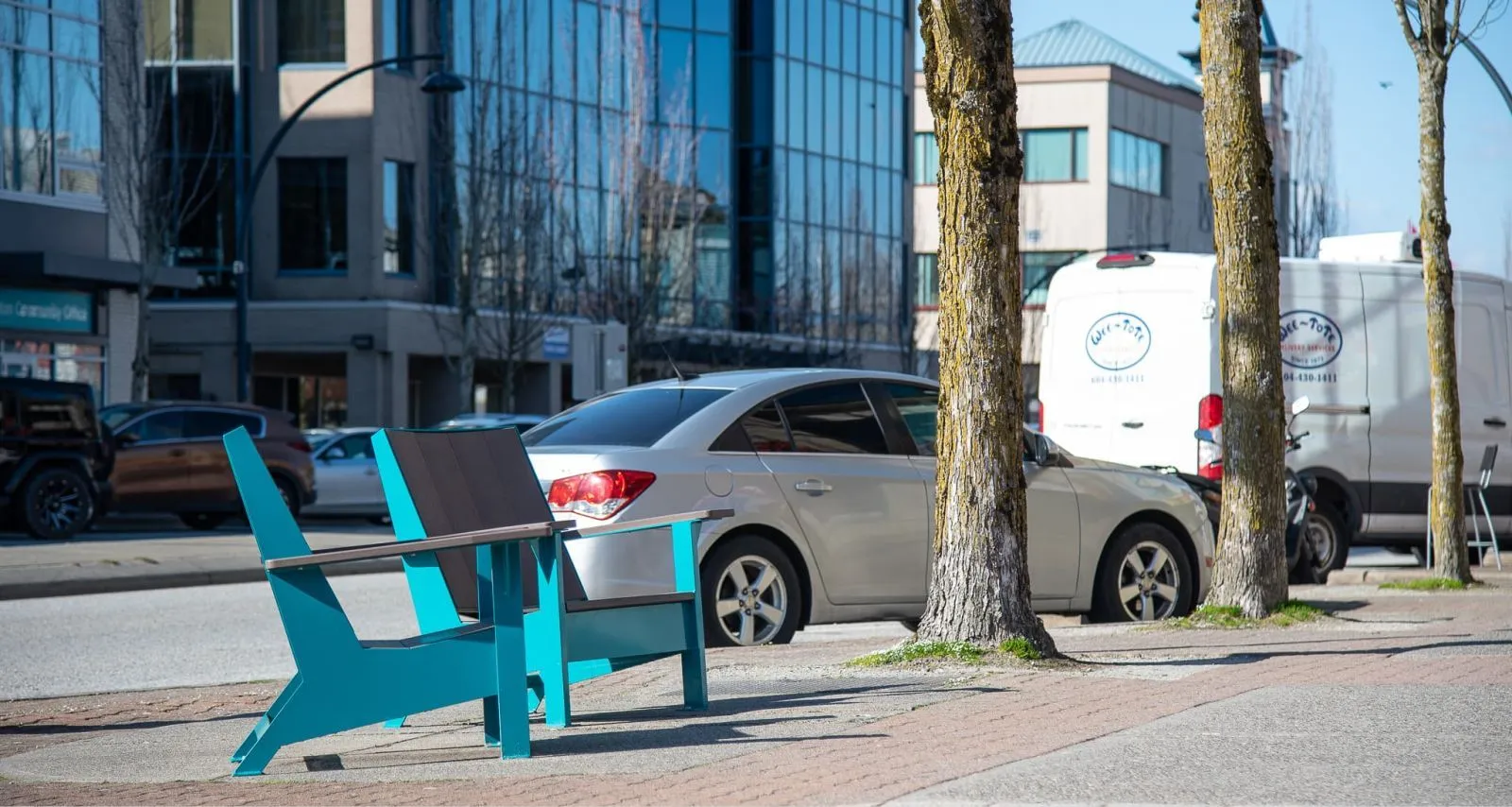 A JEM Lounge Chair in a vibrant blue finish adds a pop of color to the streetscape. The chair features recycled plastic lumber seat slats and a powder-coated metal frame.