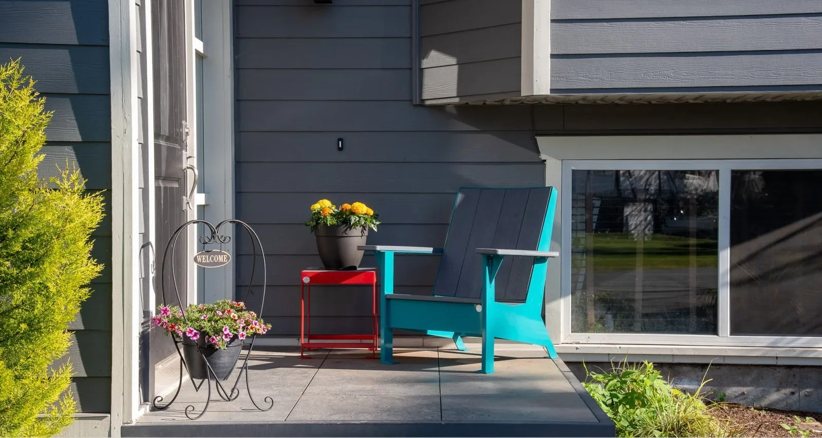 The Jem Lounge Chair and Side Table create a welcoming porch setting. The chair features Harbour Grey recycled plastic lumber and Water Blue powder coat, while the table has a Flame Red powder-coated frame.