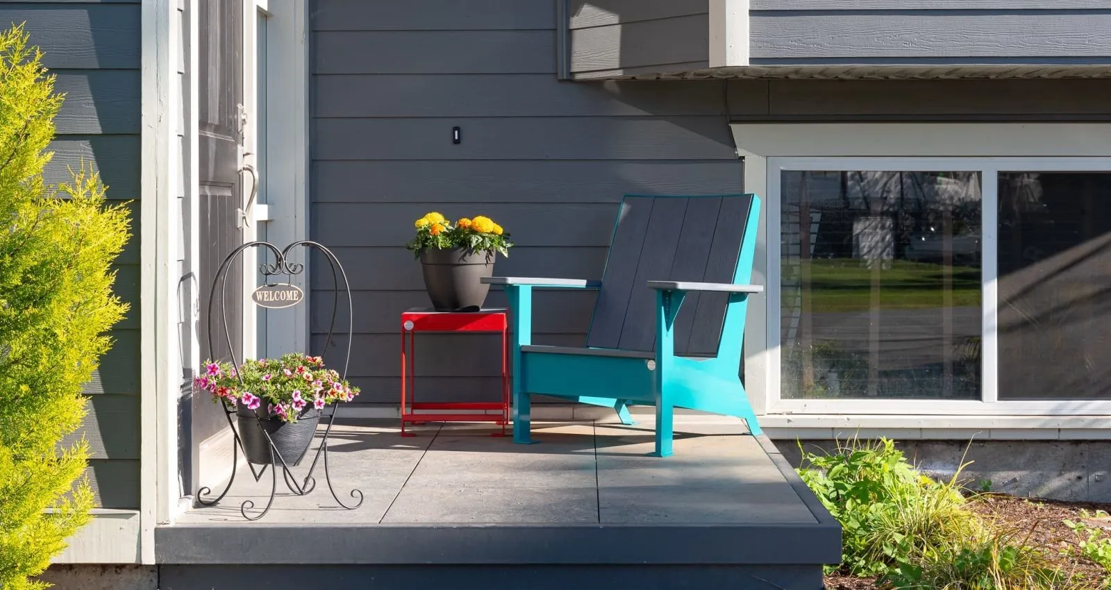 JEM Lounge Chair and Side Table on a residential porch. The chair features grey recycled plastic lumber and a custom water blue powder coat, while the table has a red powder-coated frame.