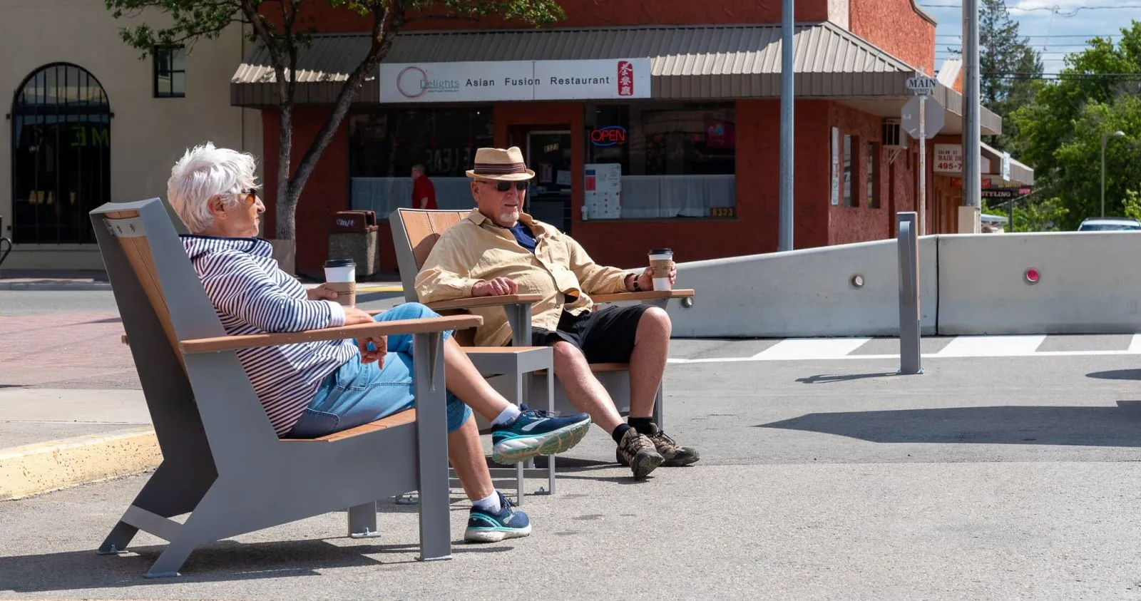 Two JEM Lounge Chairs provide seating in a downtown area. The chairs feature grey frames and natural wood-toned lumber.