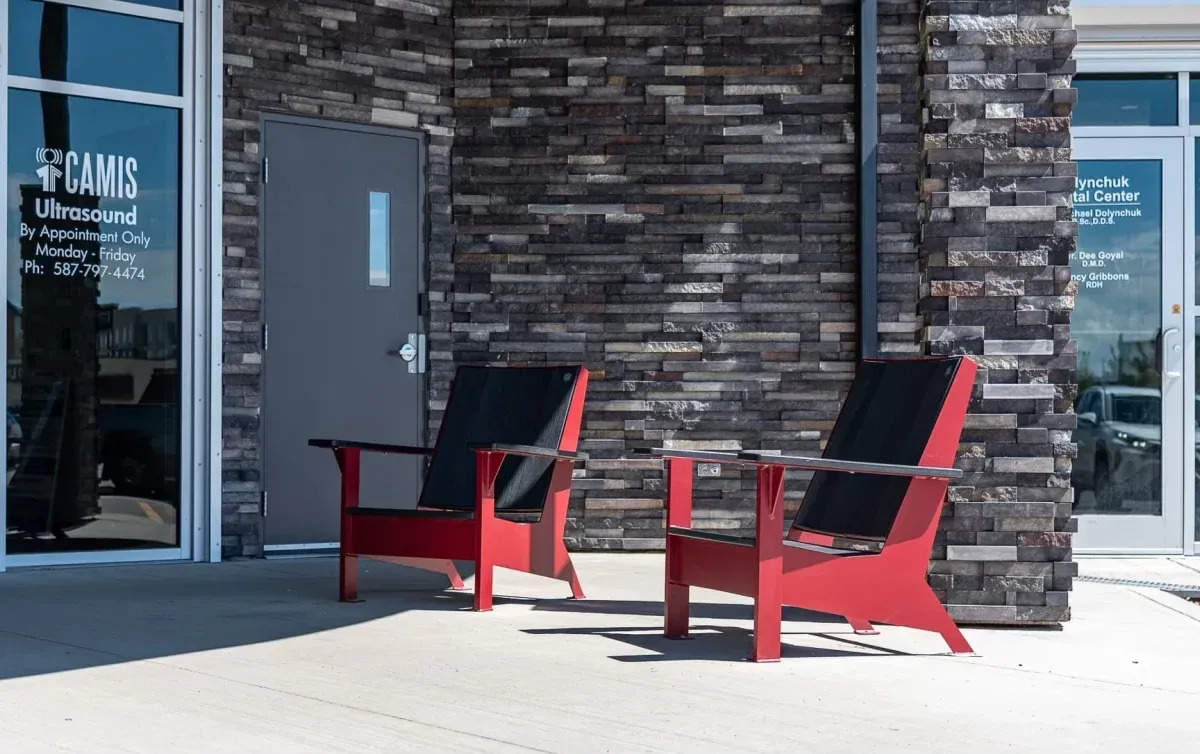 Two JEM Lounge Chairs provide seating outside a medical building. The chairs feature red frames and black woven seats.