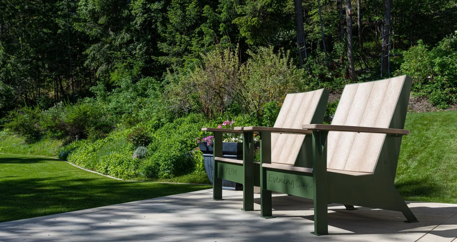 Two lounge chairs sit on a concrete patio surrounded by lush greenery. The chairs feature sand-colored recycled plastic lumber and modern khaki powder-coated frames.