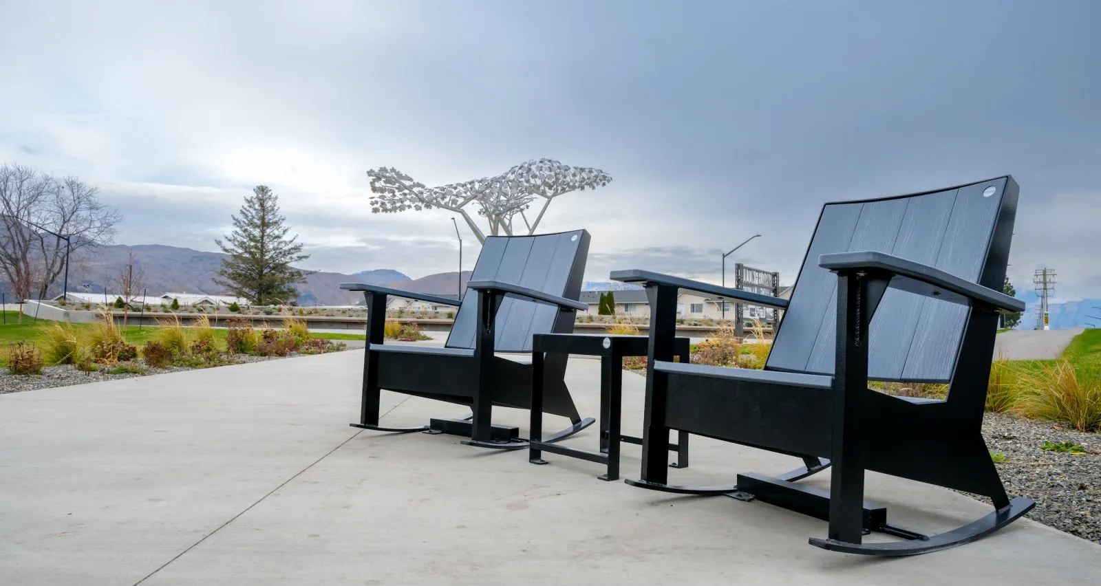 Two oversized rocking chairs and a side table create a whimsical seating area. The furniture is made with recycled plastic lumber and powder-coated metal, set on a concrete pad in a park-like setting.