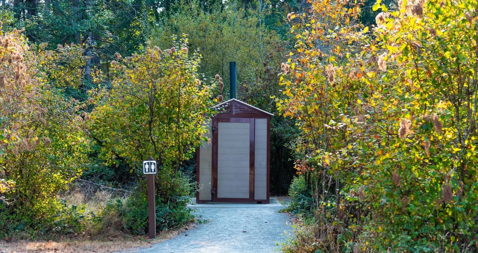 A jumbo outhouse building is nestled among lush greenery at Miracle Beach Provincial Park in Black Creek, British Columbia. The structure features light brown recycled plastic lumber and dark brown trim.