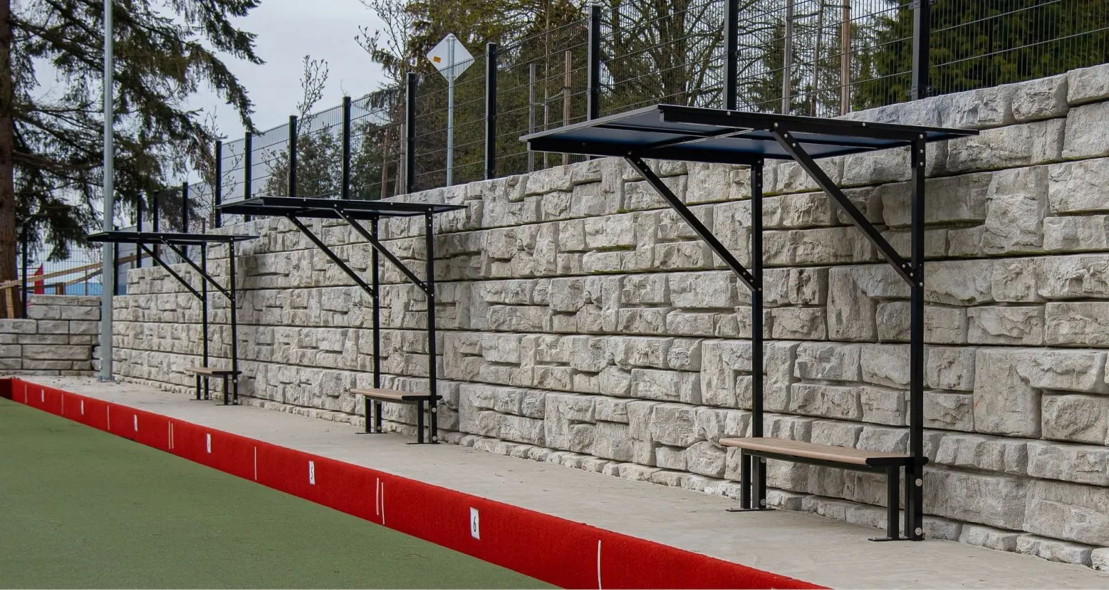 Backless benches with sun shades provide seating at the North Vancouver Lawn Bowling Club. The benches feature a simple design and are installed along a retaining wall.