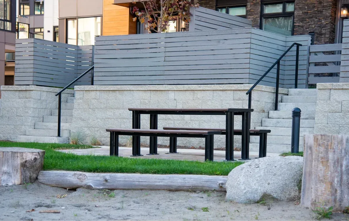 Larson picnic table with recycled plastic lumber top and black powder-coated frame, installed in a residential setting. The table sits on a grassy area near a sandy beach.
