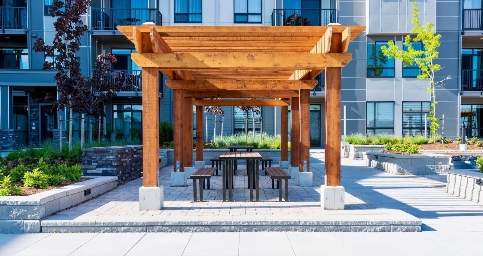 Larson picnic tables and benches provide a welcoming outdoor gathering space. The setting features a wooden pergola and modern apartment buildings in the background.