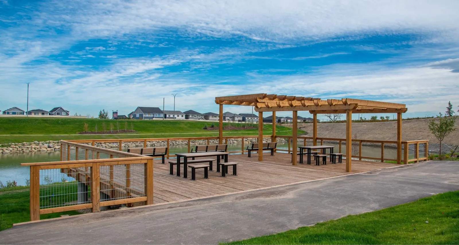 Wheelchair accessible picnic tables provide an inclusive outdoor dining space. The tables feature a black powder coat finish and are situated on a wooden deck overlooking a pond in a residential area.