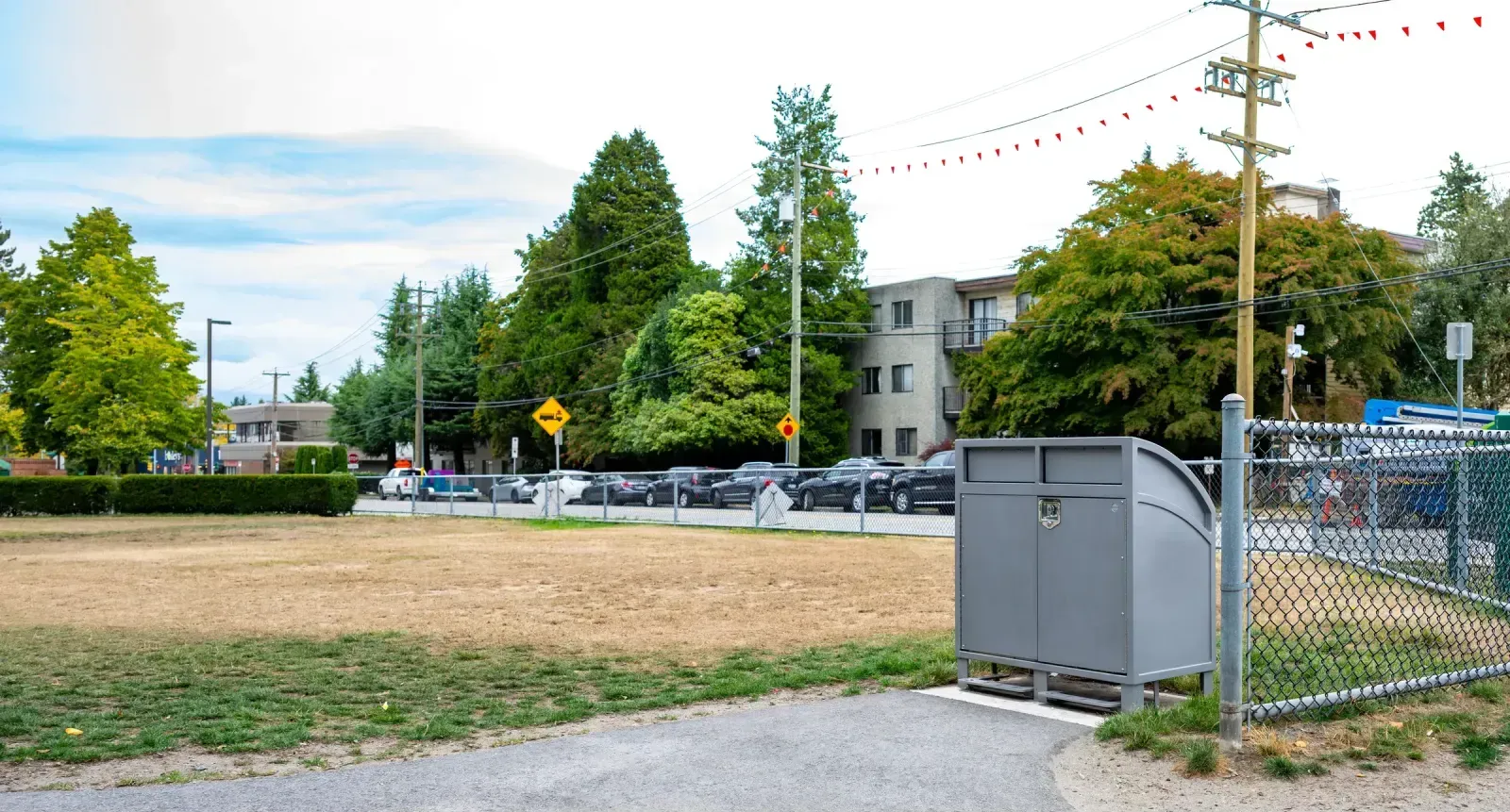 Double recycling station with a curved top is placed near a chain-link fence and a grassy area. This all-aluminum unit features optional foot pedals for hands-free operation.
