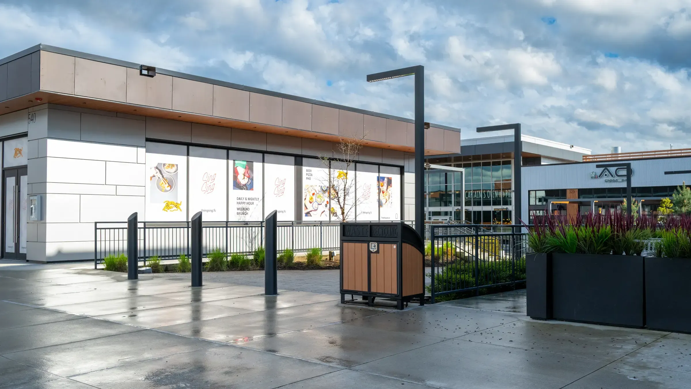 A Modena Curved Top Double Recycling Station is located outside a commercial building. The receptacle features recycled plastic lumber and a powder-coated metal frame.