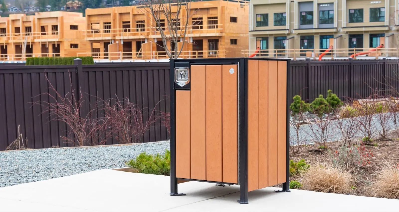 This Modena Square Top Waste Receptacle features recycled plastic lumber and a powder-coated metal frame. It is located in a residential area with new construction in the background.