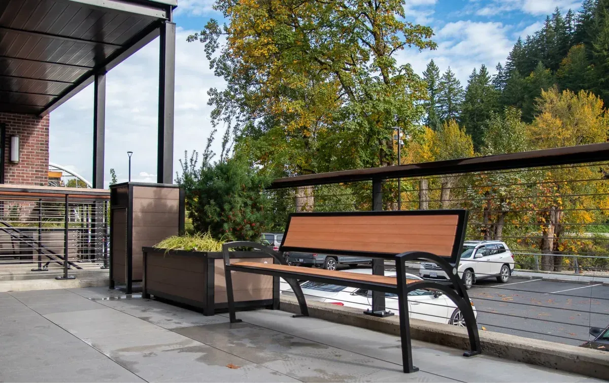 This outdoor seating area features a Modena Wide Body Bench, a planter, and a waste receptacle, all with black frames and redwood recycled plastic lumber. The setting is a commercial space in Chilliwack, British Columbia.