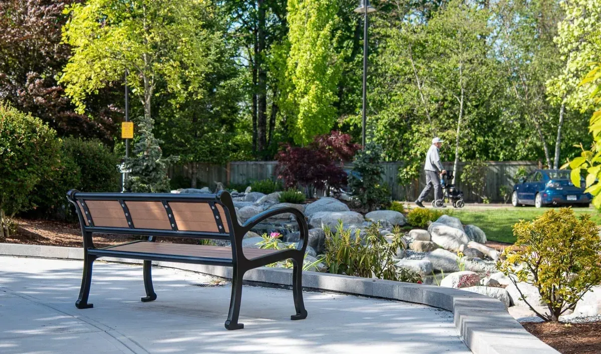 A Mountain Classic bench provides seating along a paved path in a landscaped park setting. The bench features recycled plastic lumber and a black powder-coated frame.