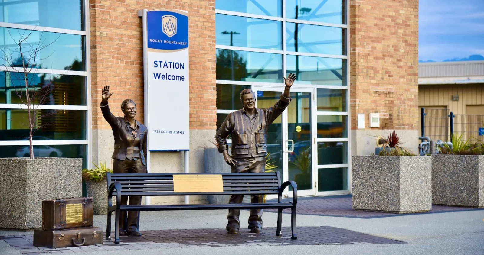 A Mountain Classic bench sits in front of the Rocky Mountaineer Station in Vancouver, BC, near a sculpture of a couple with suitcases.