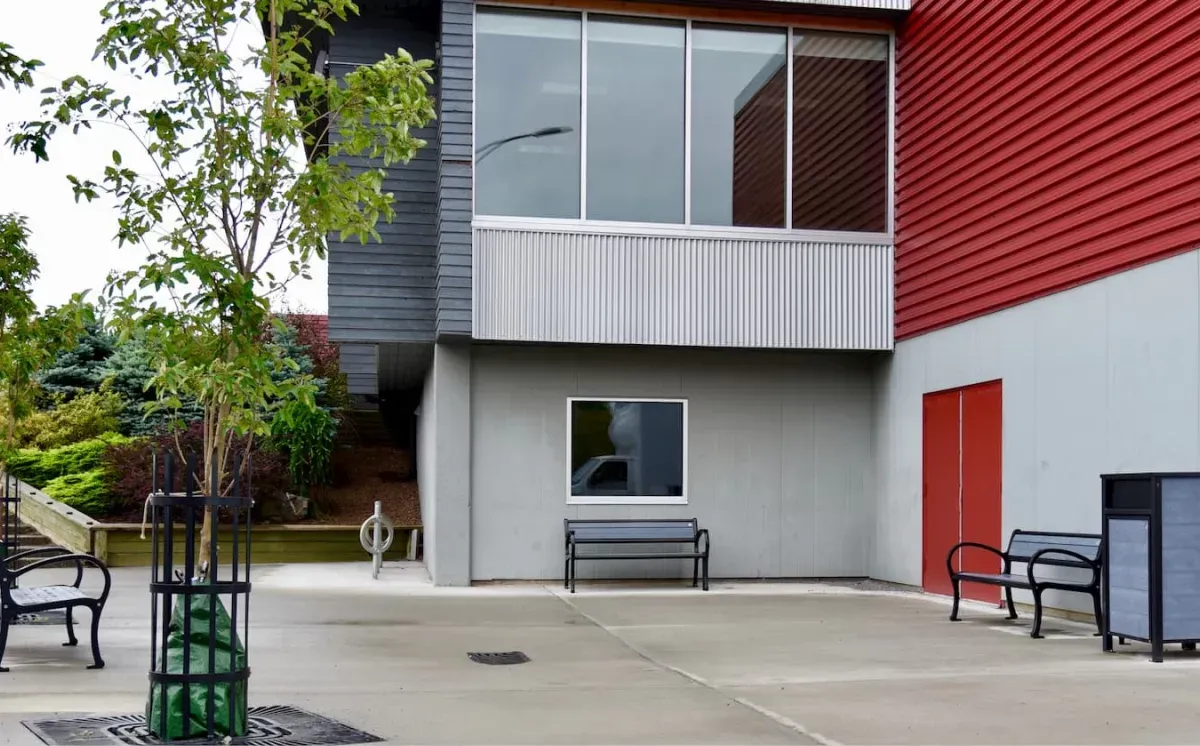 Benches provide seating in a paved outdoor area near a modern building. An Urban Form Large Capacity Waste Receptacle is also present.