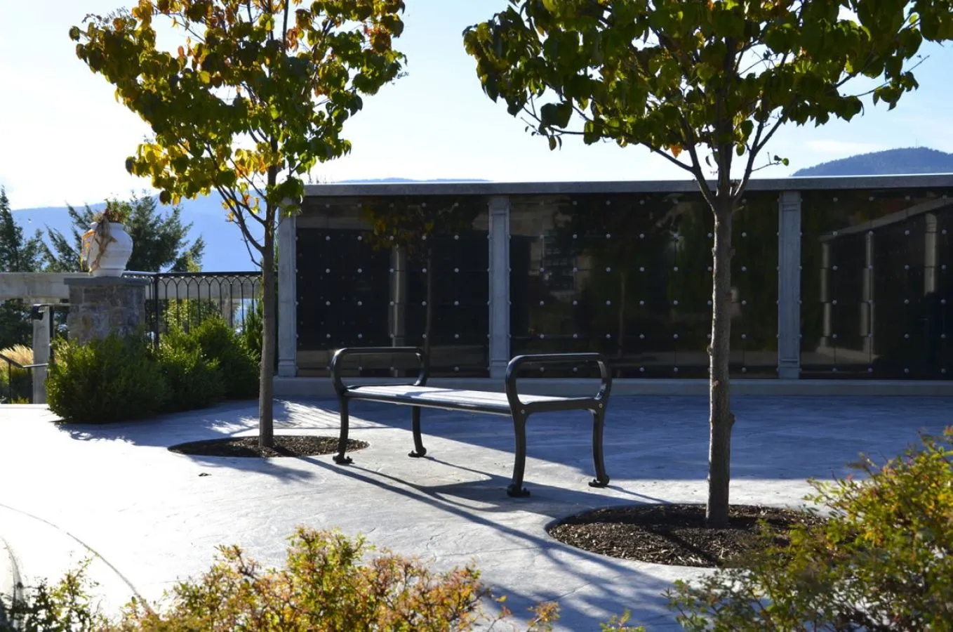A Mountain Classic Straight Bench provides seating in an outdoor space with a memorial wall and young trees. The bench offers a place for contemplation and rest in a serene setting.