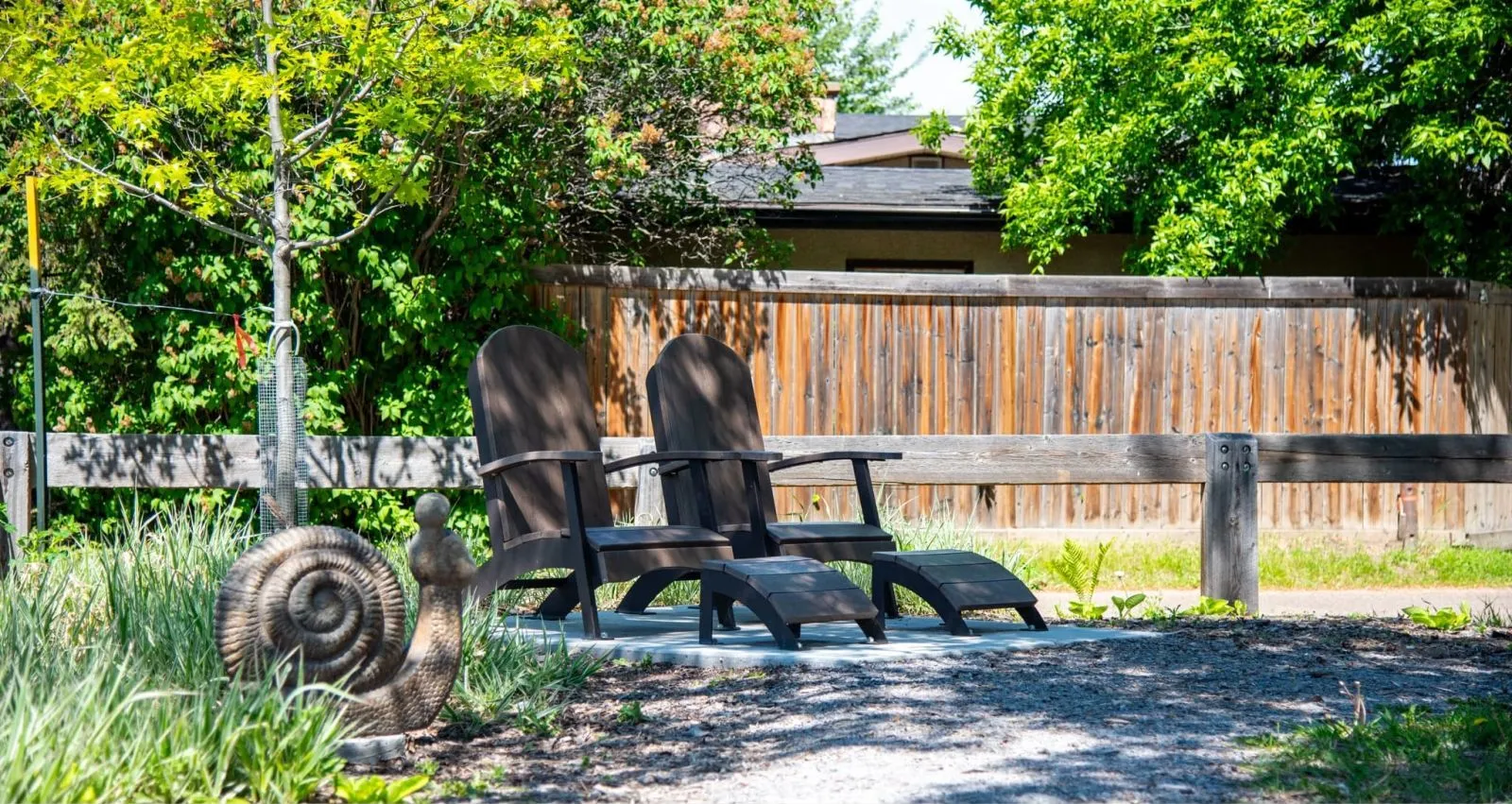 Two lounge chairs with footrests sit on a concrete pad in a garden setting. The chairs are made from recycled plastic lumber.