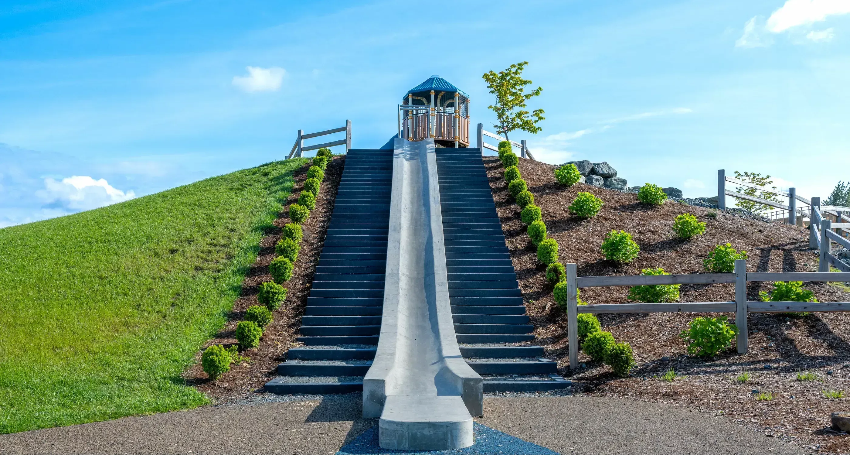 Outdoor steps with a slide provide access to a hilltop gazebo. The steps are flanked by landscaping and a wooden fence.