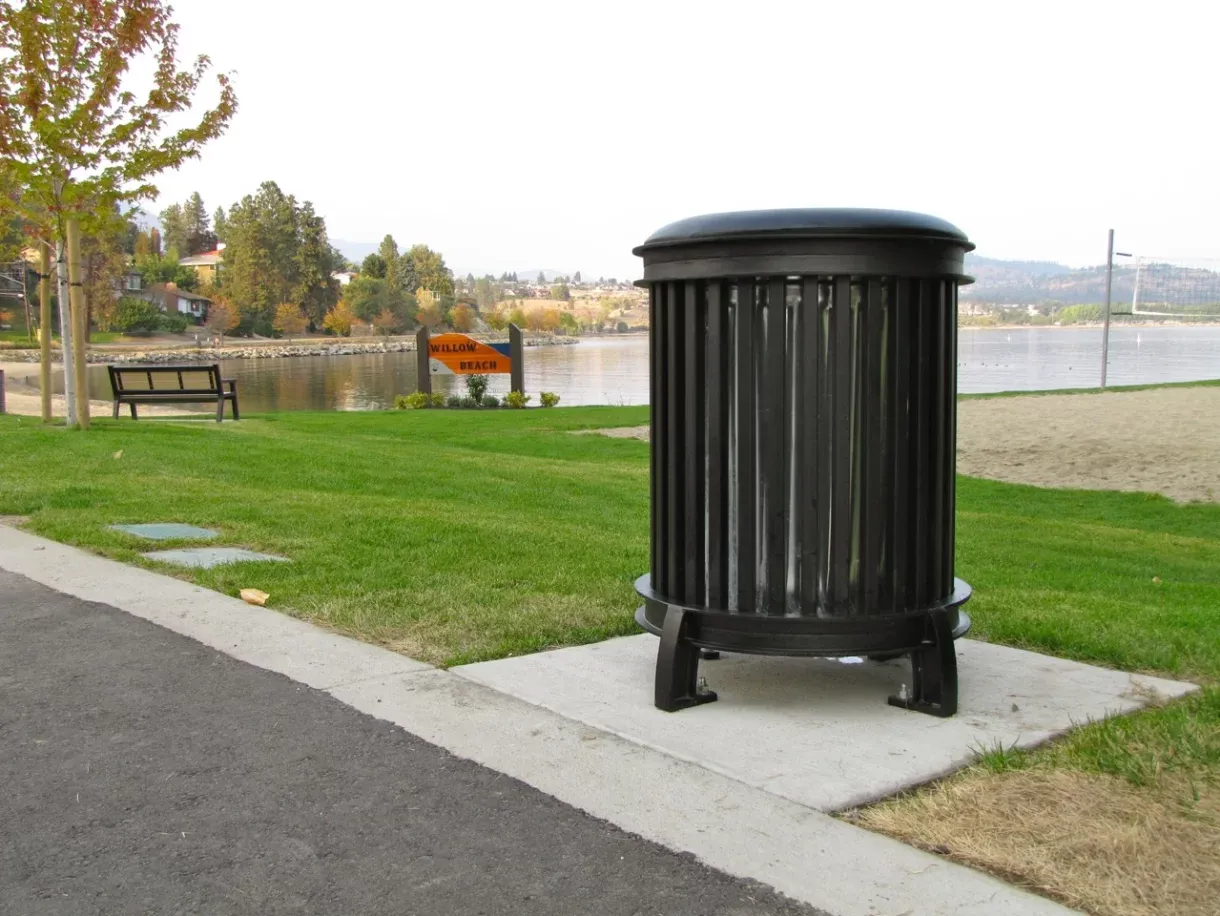 A Beselt Round Waste Receptacle sits on a concrete pad near a beach. The black powder-coated bin is placed along a paved path with a view of Willow Beach.