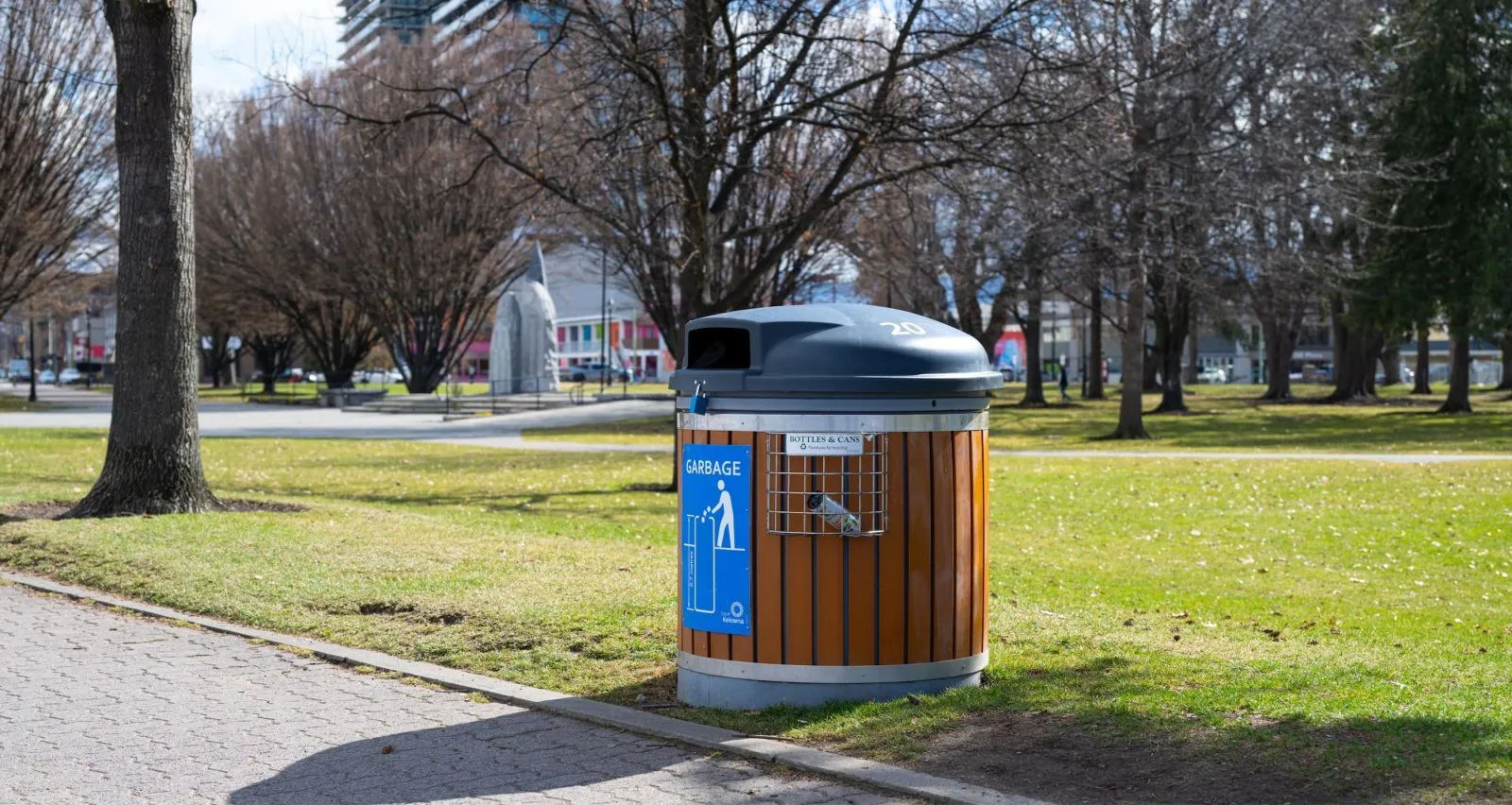 A round bottle and can recycling bin is situated in a park setting. The bin features vertical recycled plastic lumber slats and a dark grey lid.