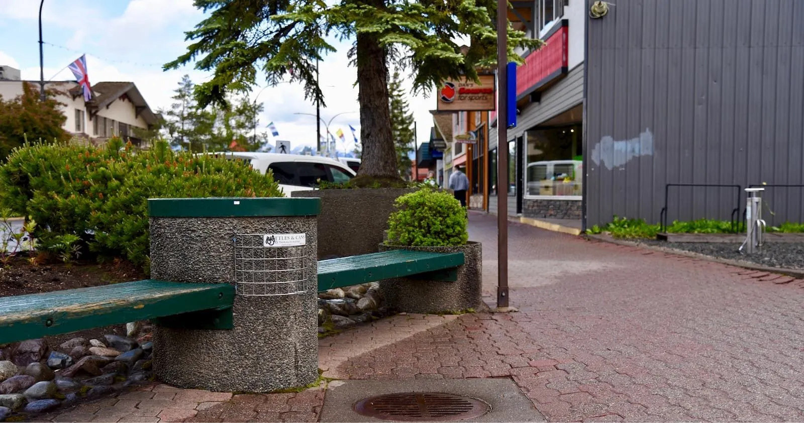 Concrete bench with a green recycled plastic lumber seat and a bottle and can recycling bin on a brick sidewalk. This site furniture provides seating and recycling options in a commercial area.