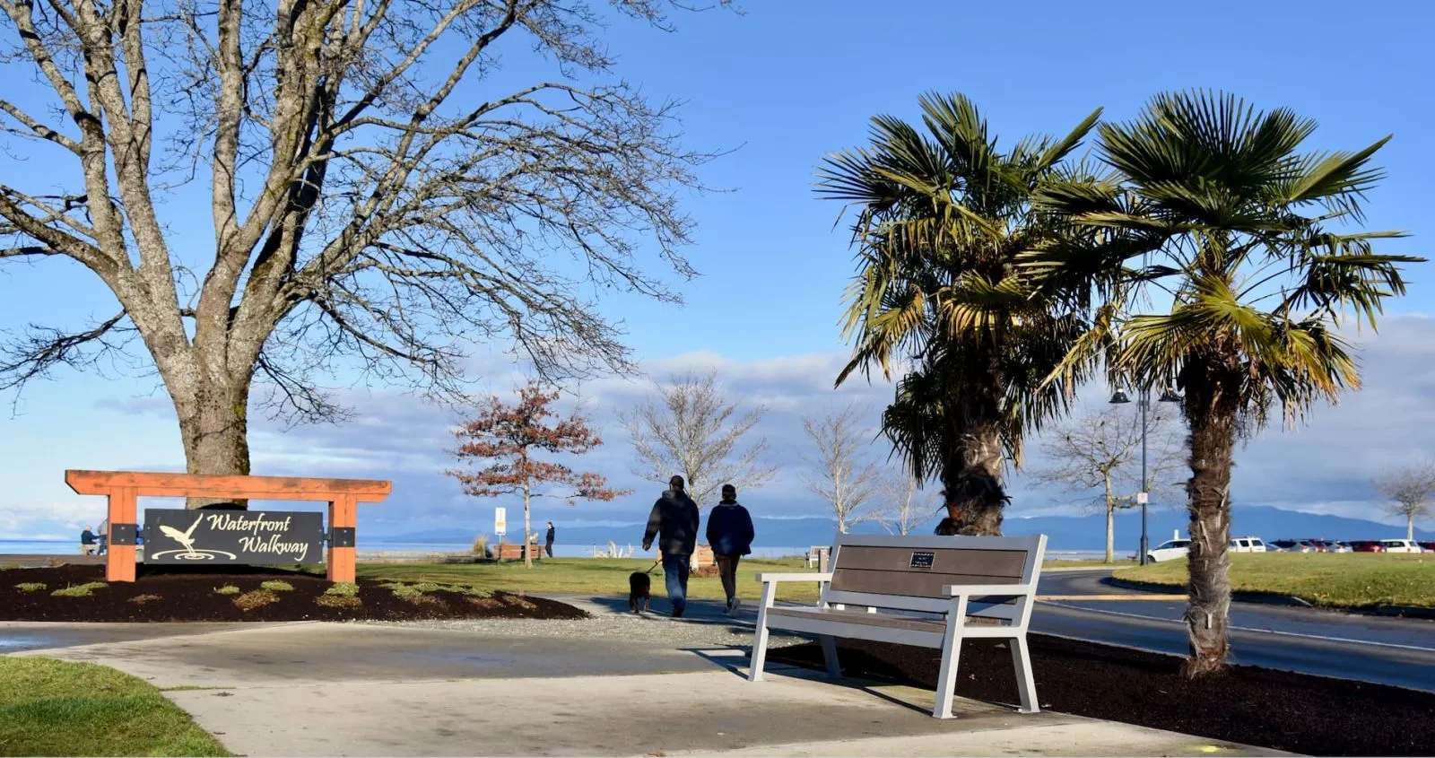 Rutherford Angled Leg bench with a memorial plaque sits along the Waterfront Walkway in Parksville, British Columbia. The bench provides seating along the walkway with a view of the ocean.