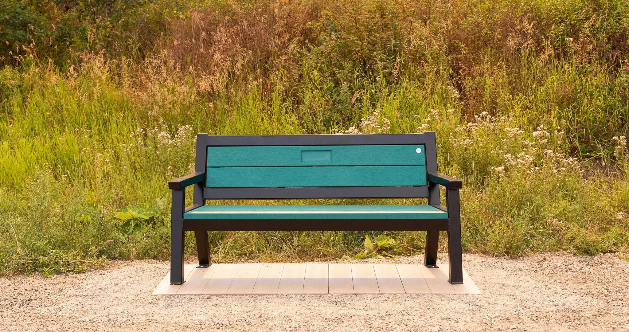 Rutherford bench with green recycled plastic lumber and a textured black frame sits on a gravel path surrounded by tall grasses. This durable and comfortable bench provides a relaxing spot in a natural setting.