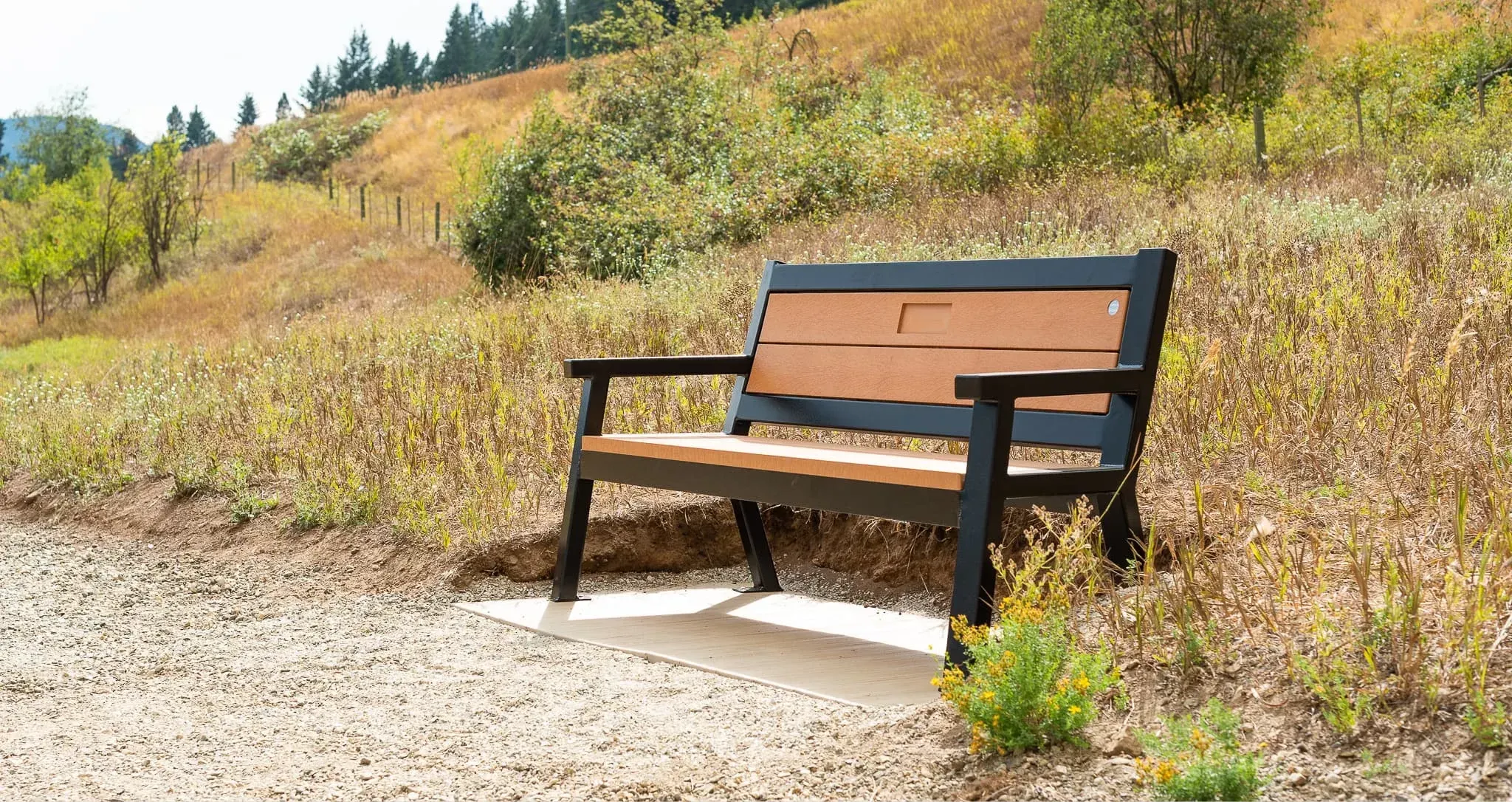 Rutherford bench with redwood recycled plastic lumber and textured black frame, installed on a gravel path. The bench provides seating along the Salmon Trail in Lumby, British Columbia.