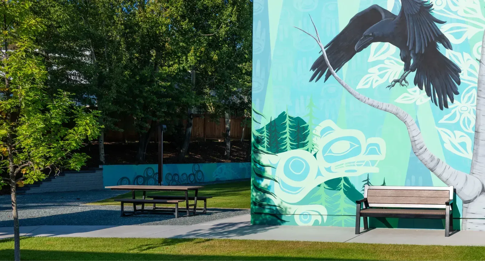 Outdoor furniture including a Rutherford picnic table and bench are placed in a park setting next to a mural. The picnic table features a natural wood finish, complementing the natural environment.