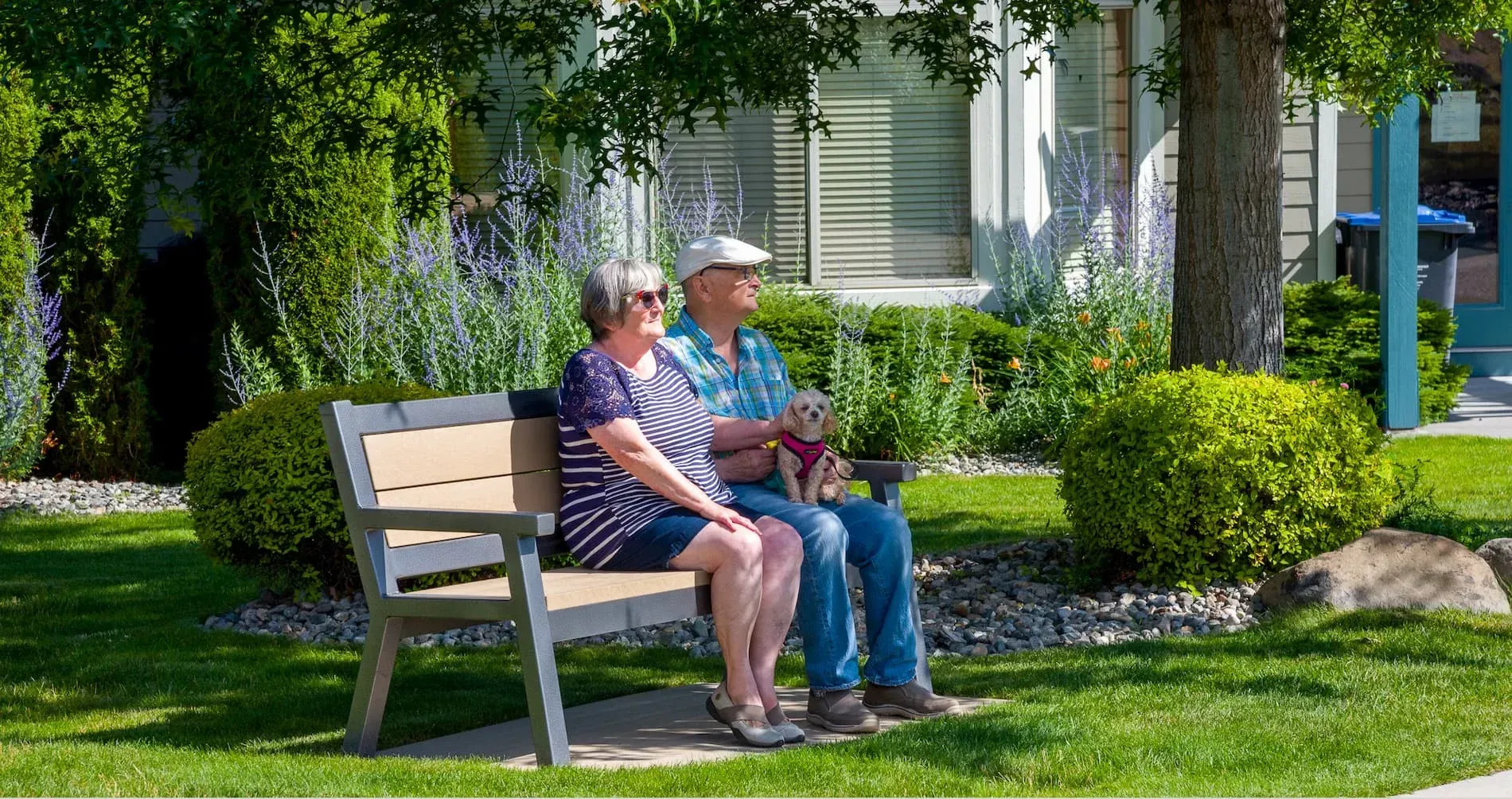Rutherford bench provides comfortable seating in a landscaped garden setting. The bench features recycled plastic lumber slats and a powder-coated metal frame.