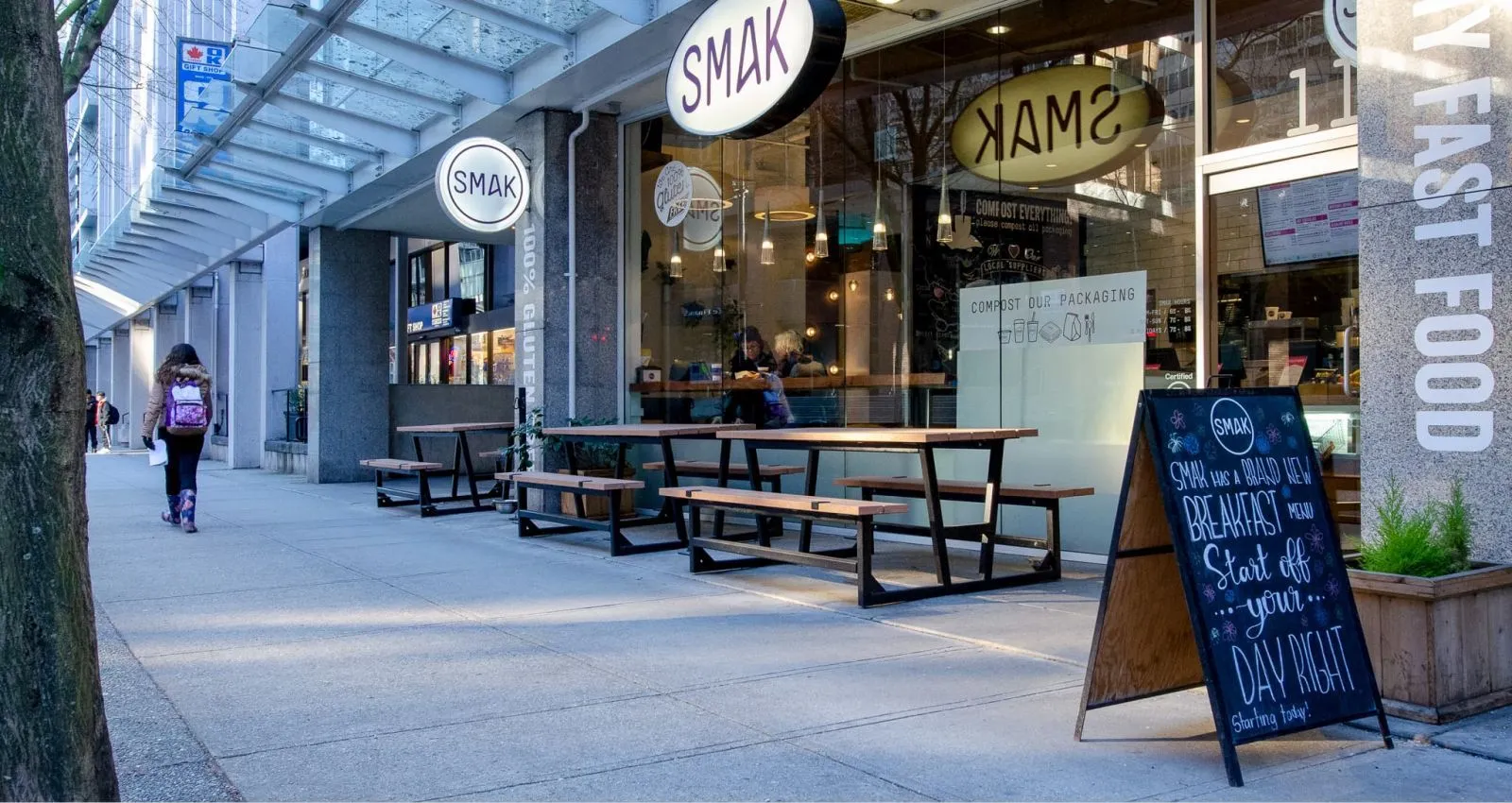 Rutherford picnic tables provide outdoor seating in front of Smak Healthy Fast Foods in Vancouver, British Columbia. The tables feature a black metal frame and light-colored lumber.