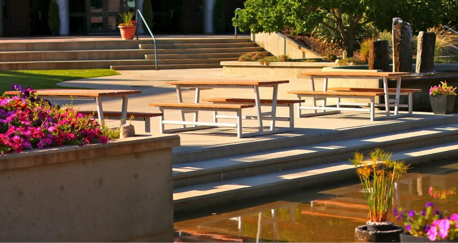 Several picnic tables provide outdoor seating near a pond and landscaped area. These tables feature light-colored tops and silver frames.