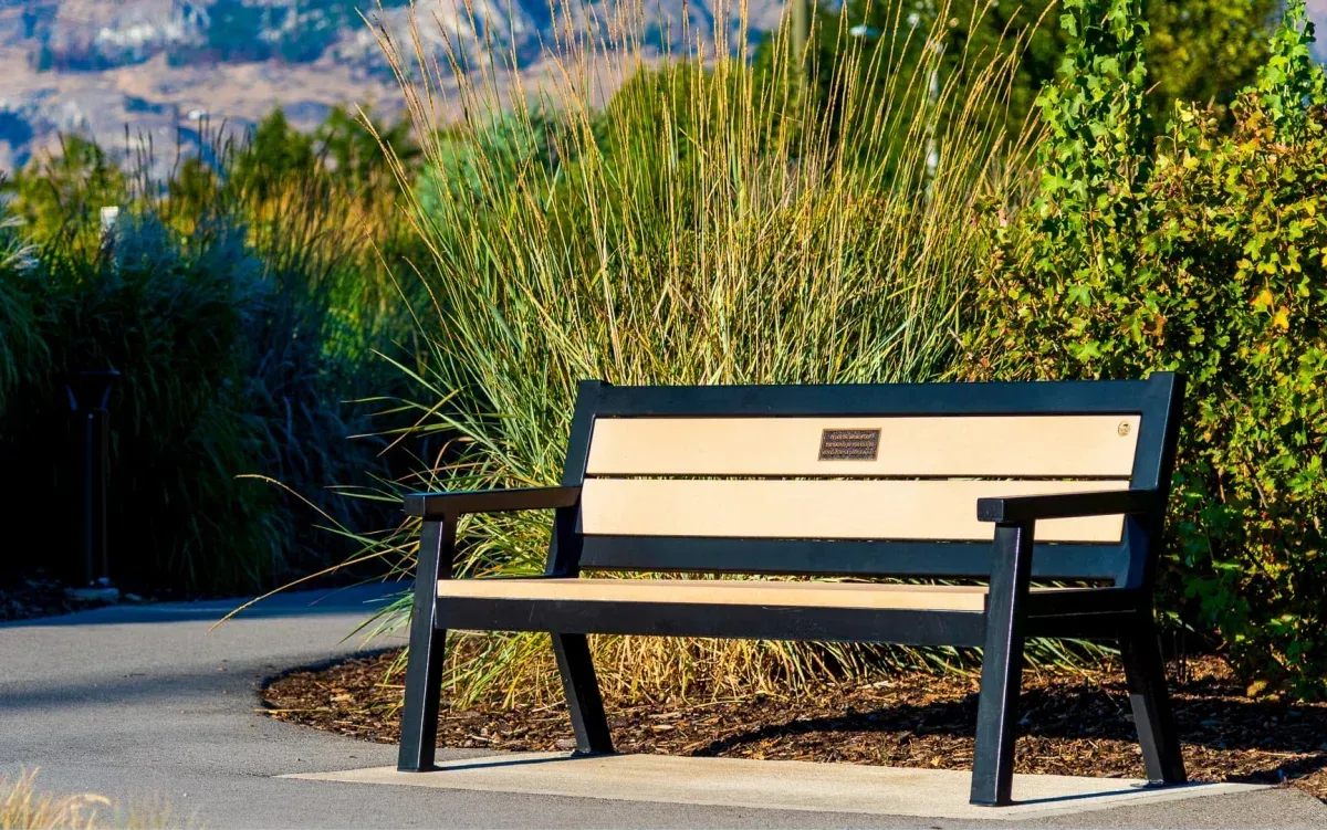 A Rutherford bench sits along a paved path surrounded by lush greenery. The bench features recycled plastic lumber and a powder-coated metal frame, offering a comfortable and durable seating option in an outdoor setting.