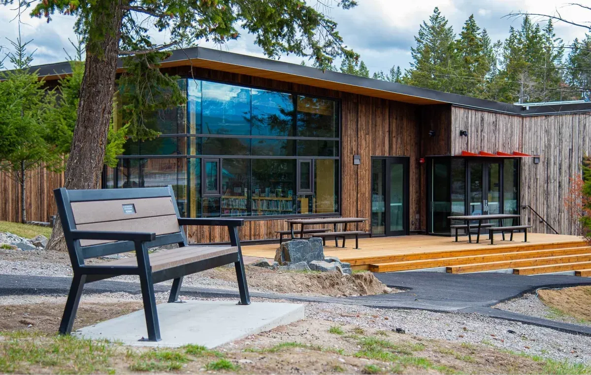 A Rutherford bench provides seating outside a modern building with wood siding and large windows. Picnic tables are visible on the wooden deck.