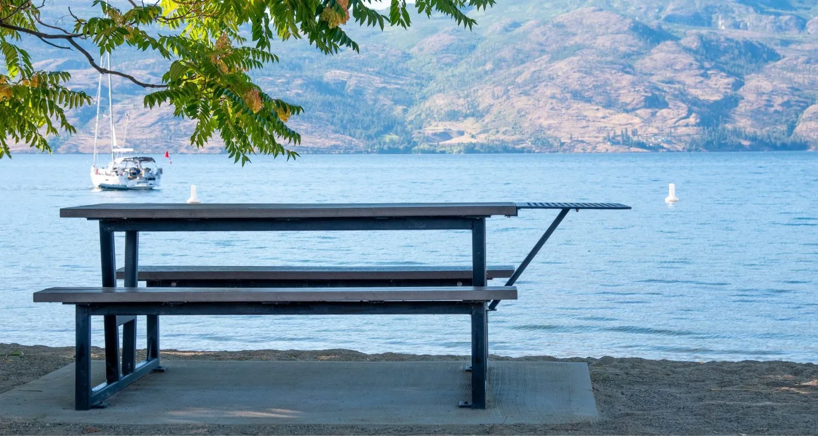 A Rutherford picnic table with a BBQ extension sits on a concrete pad near the beach. The table features recycled plastic lumber and a powder-coated metal frame, offering a durable and stylish outdoor seating solution.