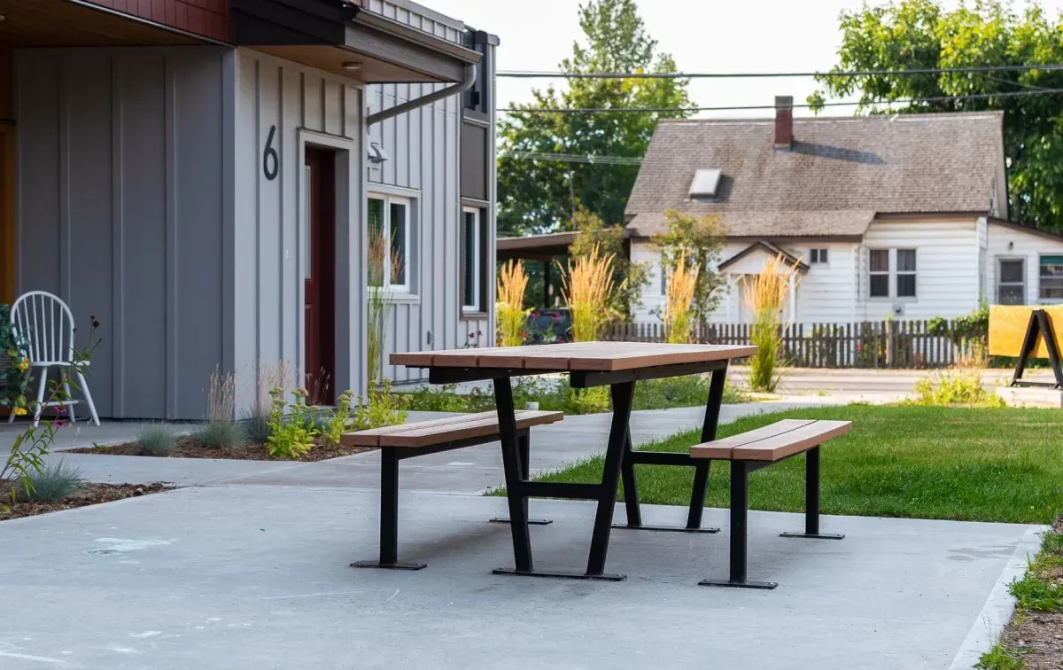 Wheelchair-accessible picnic table with a brown top and black frame sits on a concrete patio. The table is located outside a building with light gray siding and a red door.