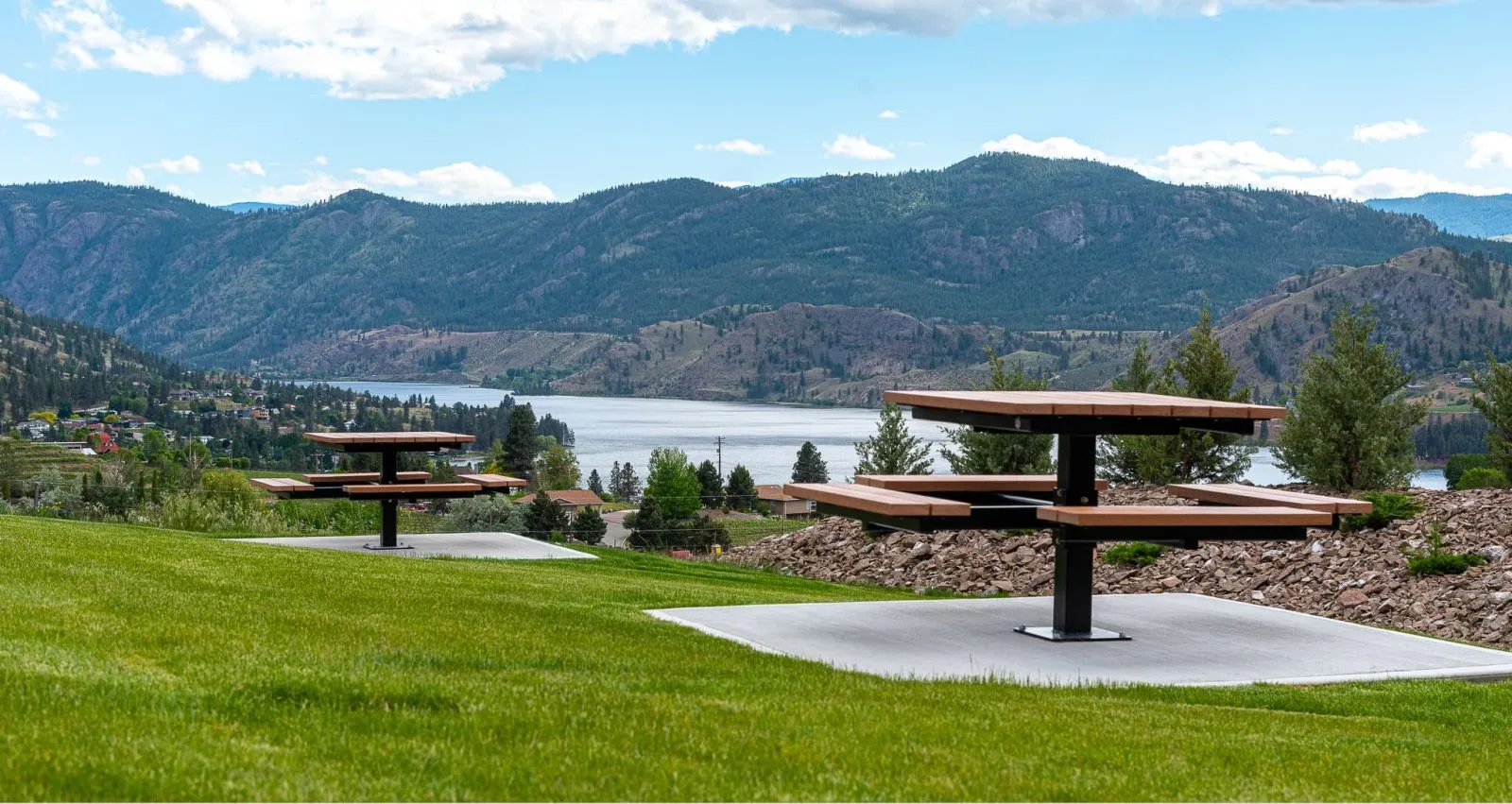 Two single pedestal picnic tables are installed on concrete pads in a grassy park overlooking a lake and mountains. These accessible tables provide a comfortable and stylish place to enjoy the scenic views.