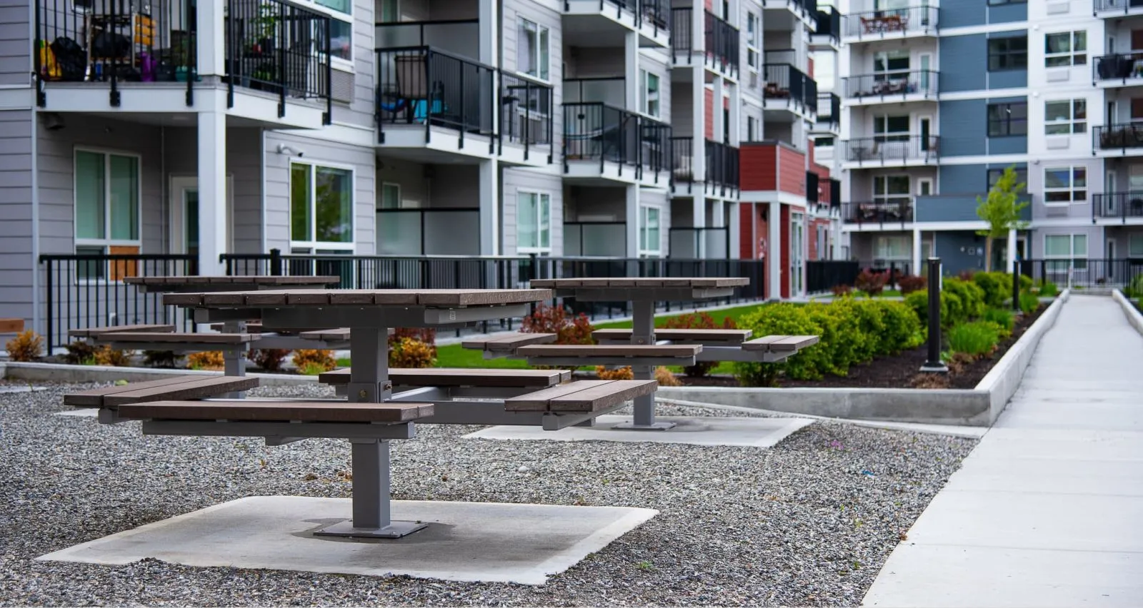 These single pedestal picnic tables offer a comfortable and accessible outdoor dining solution. The tables are set in a gravel courtyard surrounded by modern apartment buildings and landscaping.