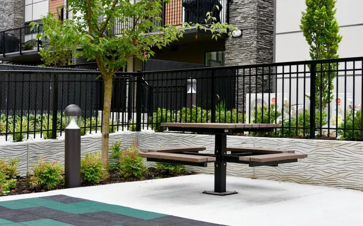 Single pedestal picnic table in an outdoor courtyard setting with modern architecture and landscaping. The table features recycled plastic lumber and a powder-coated metal frame.